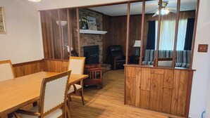 Una mesa de comedor de madera con sillas, una chimenea, un televisor y un sofá a rayas en una habitación con paneles de madera y un ventilador de techo.