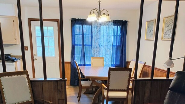 Un comedor con una mesa y sillas de madera, una araña y vistas al exterior a través de una ventana con cortinas azules.