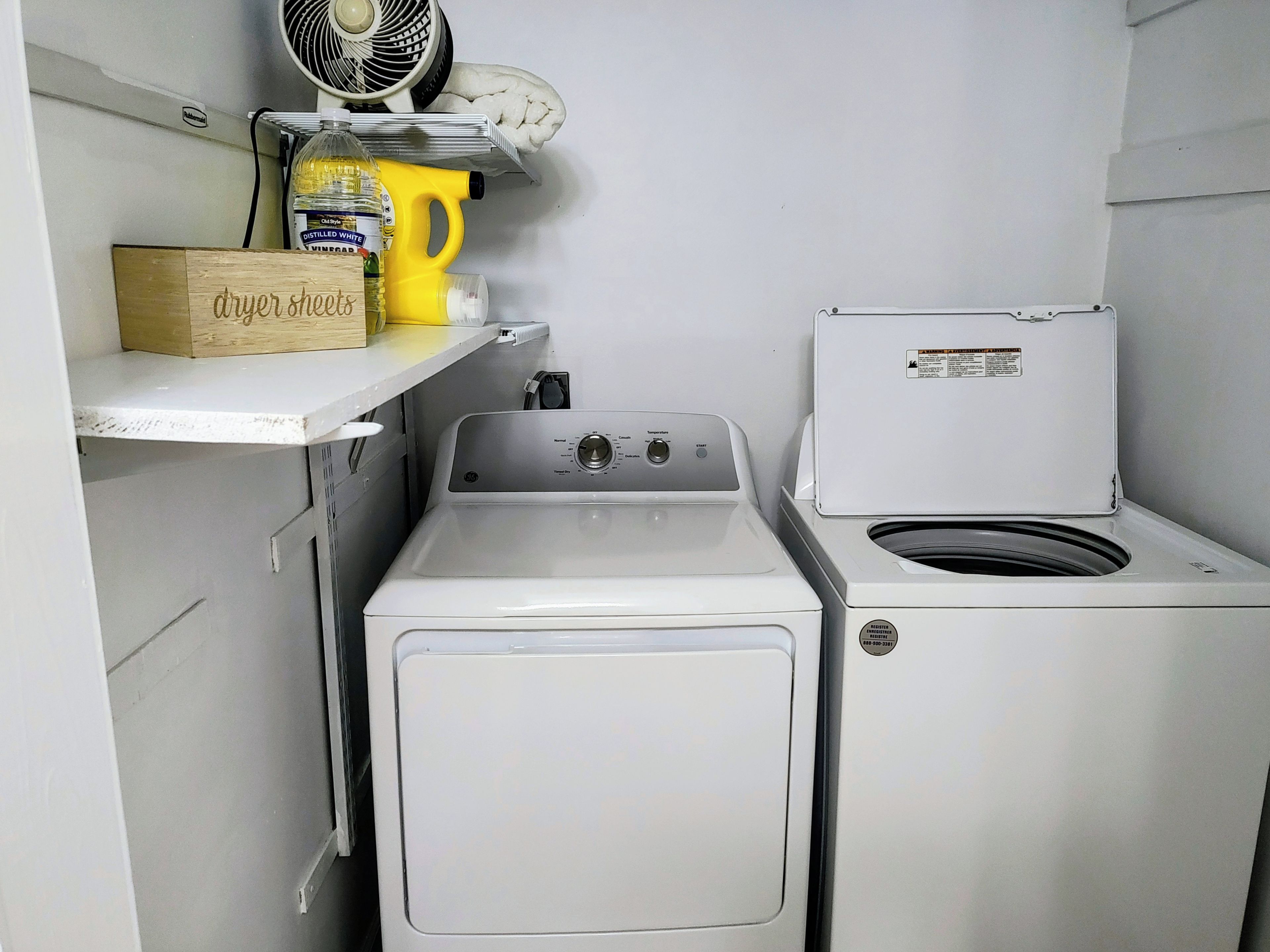 Un espace réduit dédié à la buanderie, comprenant un lave-linge et un sèche-linge à chargement frontal, une étagère avec une boîte étiquetée « feuilles pour sèche-linge » et une bouteille de liquide vaisselle « Olde Time ».