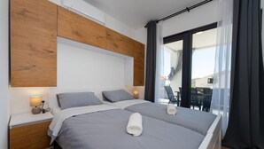 Un lit double avec du linge de lit gris et deux serviettes enroulées, un lambris en bois sur le mur, une table de chevet avec une lampe, et une vue sur l’extérieur à travers une porte vitrée.
