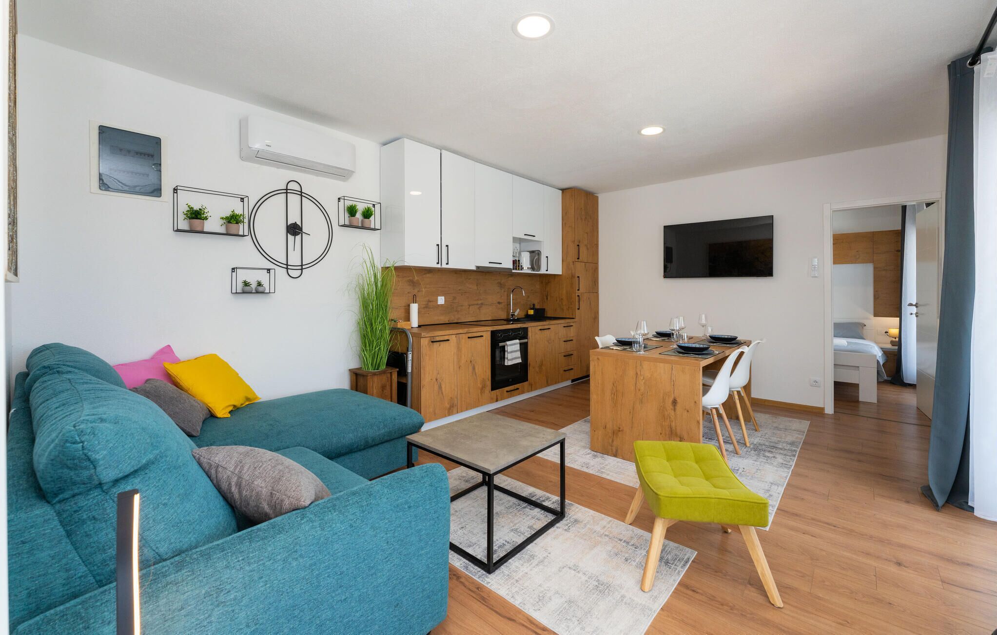 Un salon moderne avec un canapé de couleur turquoise, une table à manger en bois et un coin cuisine doté de meubles blancs et d’un plan de travail en bois.