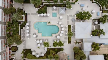 An aerial view of a hotel with a central swimming pool, multiple lounge areas with umbrellas, and surrounding greenery.