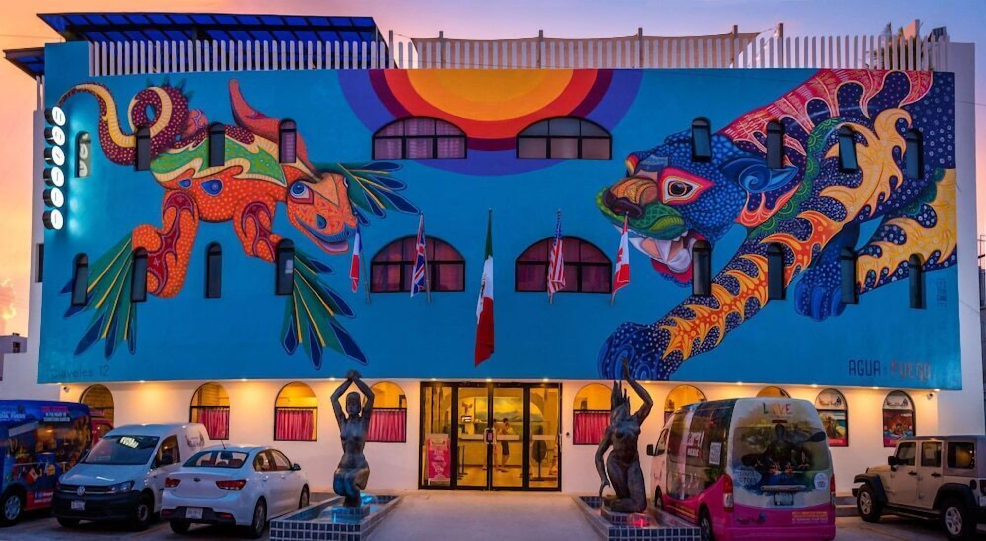 A building with a large mural of colorful animals and flags. There are cars parked in front, and a statue of a woman is visible.
