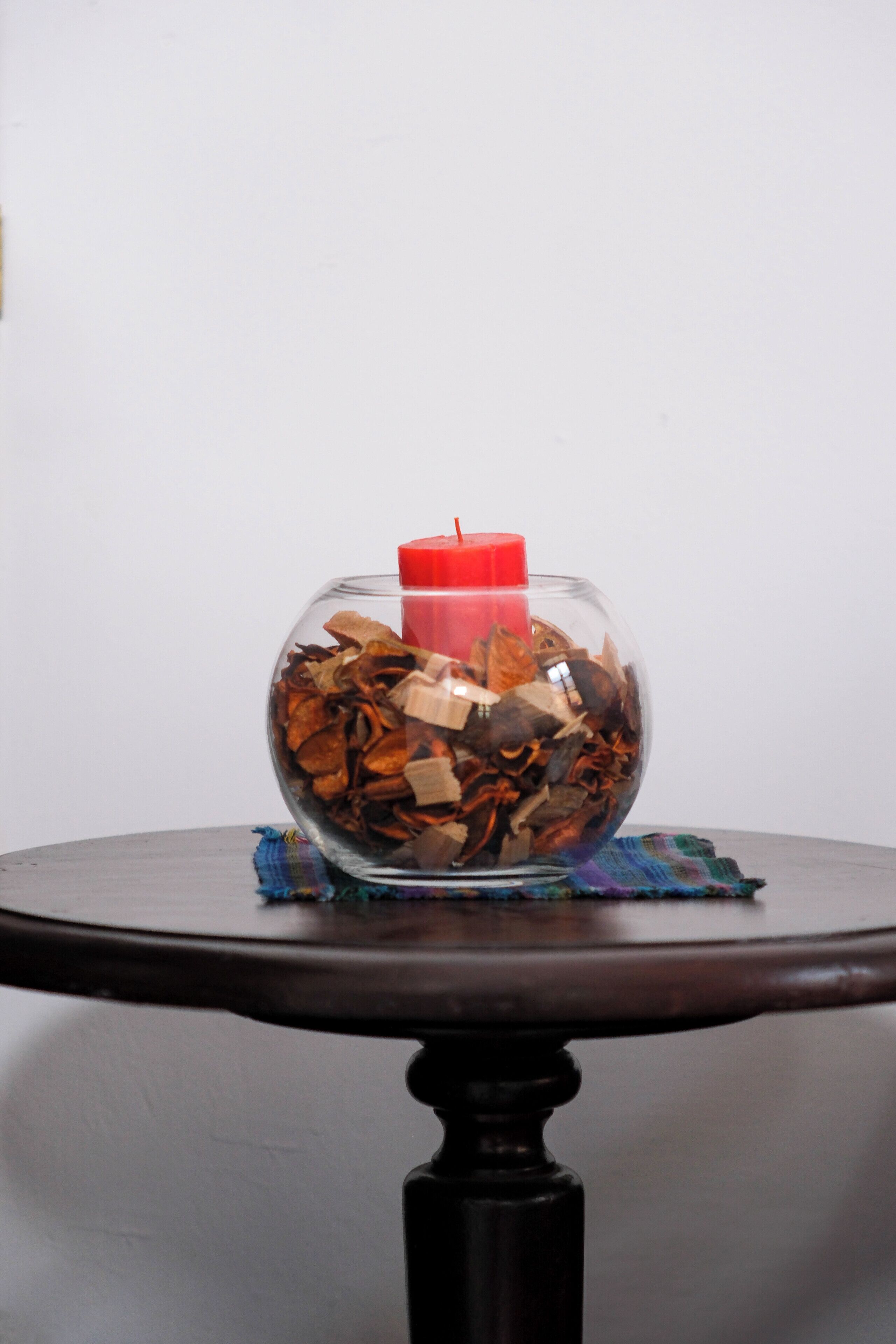 A clear glass bowl filled with various dried leaves and a red candle on a round table with a dark wooden base.