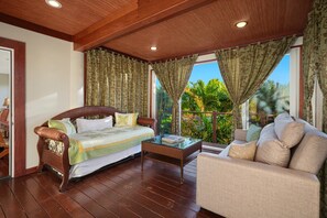 Une pièce avec un plafond en bois, un lit recouvert d’une courtepointe verte et blanche, un canapé, une table basse en verre et une vue sur une végétation luxuriante à l’extérieur.