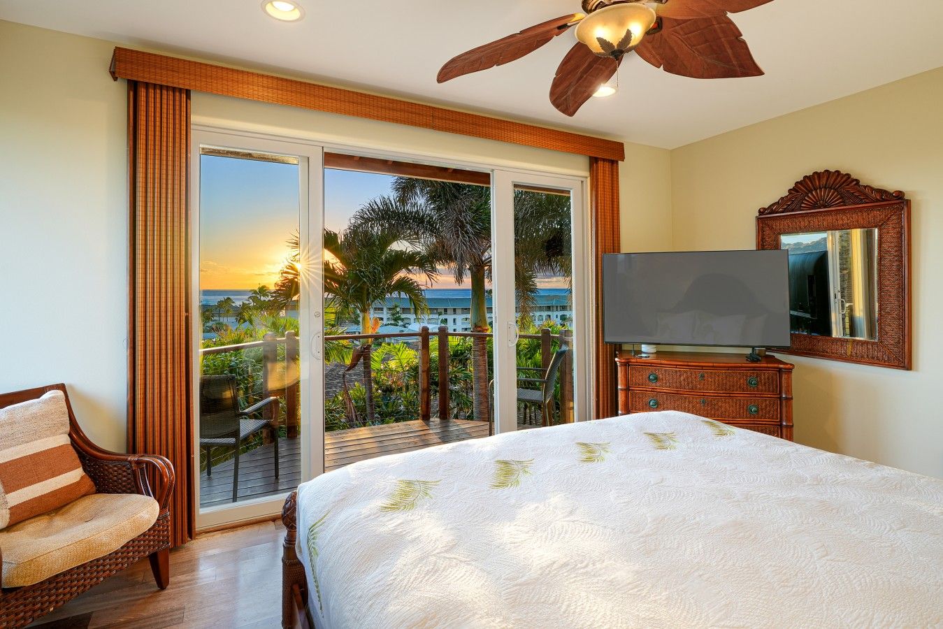 Quarto de hotel com cama, televisão, cômoda de madeira e varanda com vista para palmeiras e o oceano.