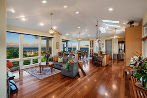 A spacious living area with a large window overlooking a scenic view, a dining area with a kitchen in the background, and a ceiling fan. - Luxe Oceanview Getaway w/ Heated Pool & Epic Sunrise (As Seen on TV) (Koloa)