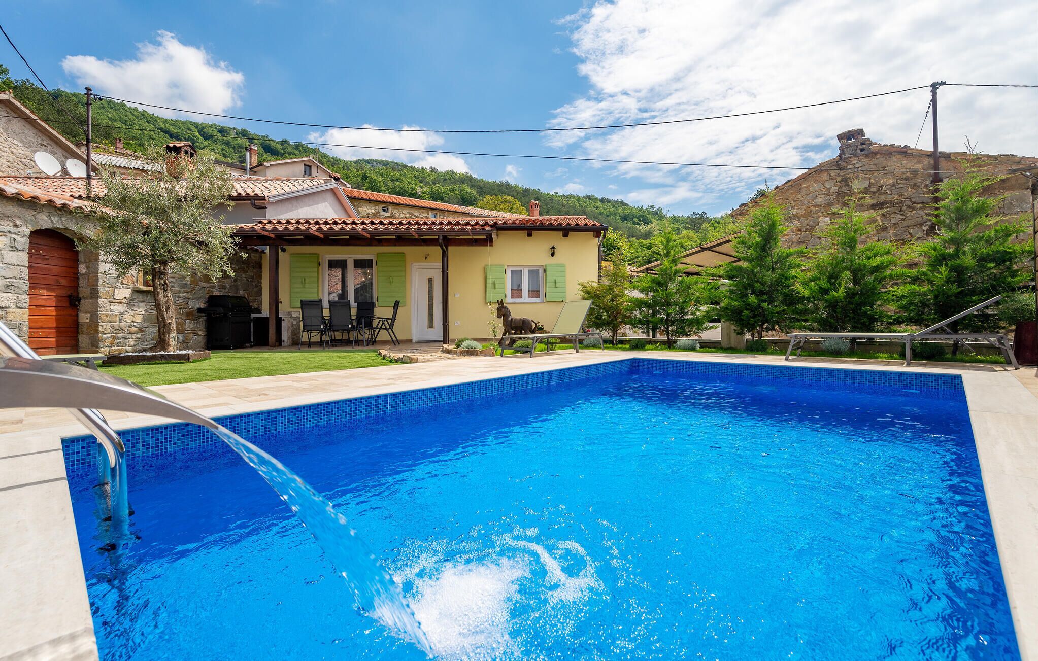 A swimming pool with a clear blue water, a stone house with a tiled roof, and a green lawn.