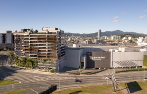A modern building with a large sign for 'BALNEARIO Shopping' and a view of a cityscape with mountains in the background. - The Spot One - Luxo e acesso direto ao Balneário Shopping (Balneário Camboriú)