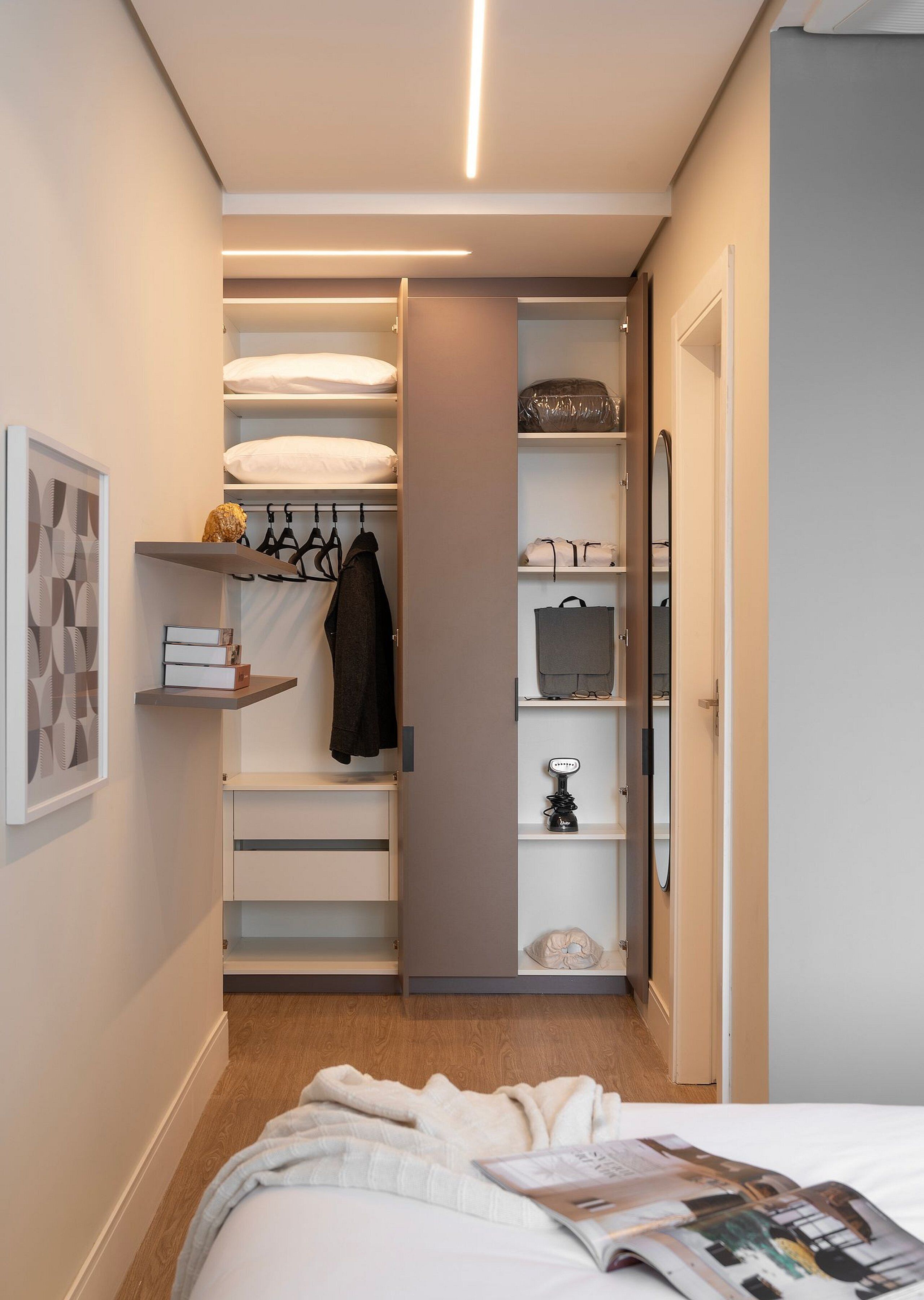 A bedroom with a neatly made bed, a bedside table, a closet with hanging clothes, and a shelf with a lamp.