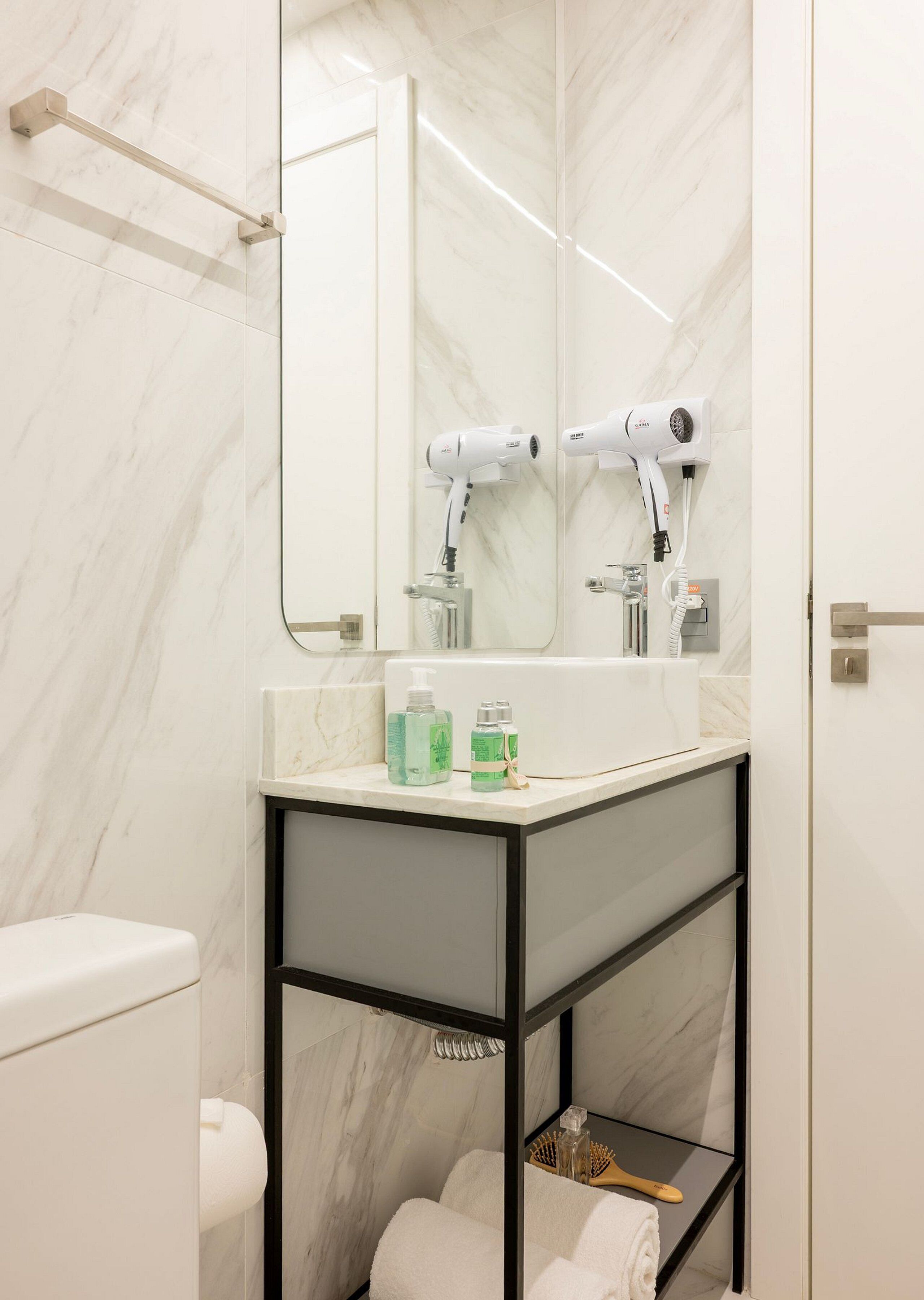 A bathroom with a marble wall, a white sink, a mirror, two hair dryers, and a towel rack.