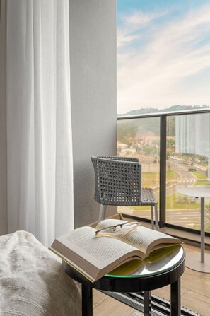 Un balcón con vista a un paisaje urbano, una silla de mimbre, una mesa y un libro abierto sobre una mesa de vidrio.