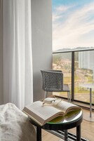 Uma varanda com vista para o horizonte urbano, uma cadeira de vime, uma mesa e um livro aberto sobre uma mesa de vidro.
