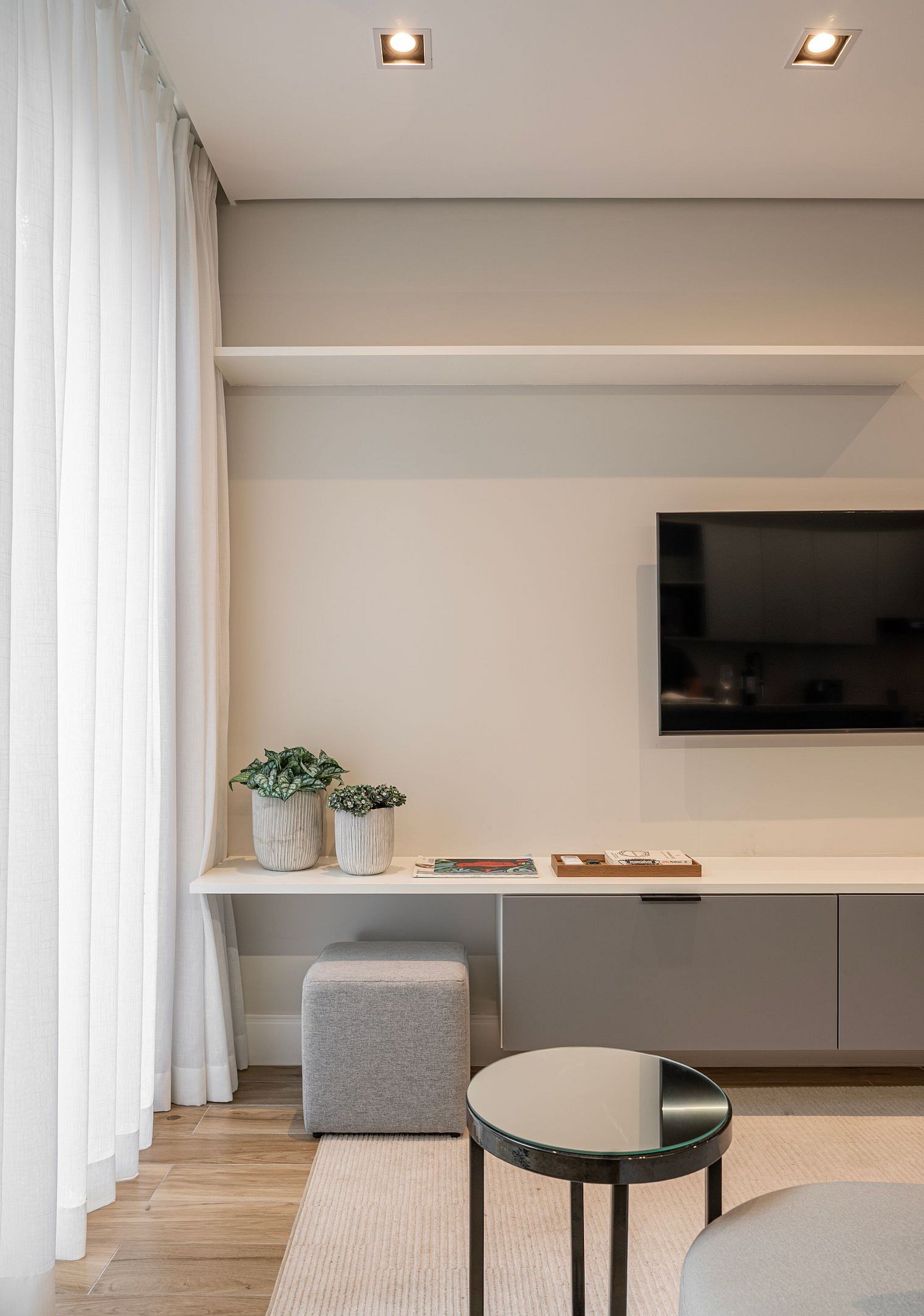 Un salon moderne avec un téléviseur à écran plat, une console blanche, deux plantes en pot, un pouf gris et une table ronde en verre.