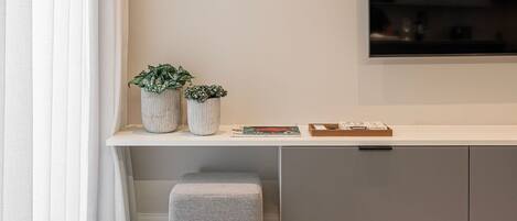 Un salon moderne avec un téléviseur à écran plat, une console blanche, deux plantes en pot, un pouf gris et une table ronde en verre.