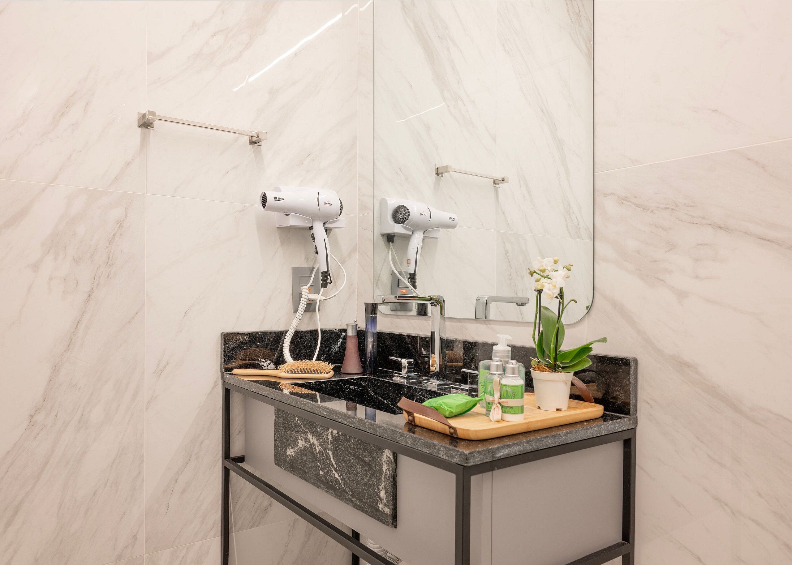 A bathroom with a marble wall, a sink with a mirror, two hair dryers, a wooden cutting board, and a potted orchid.