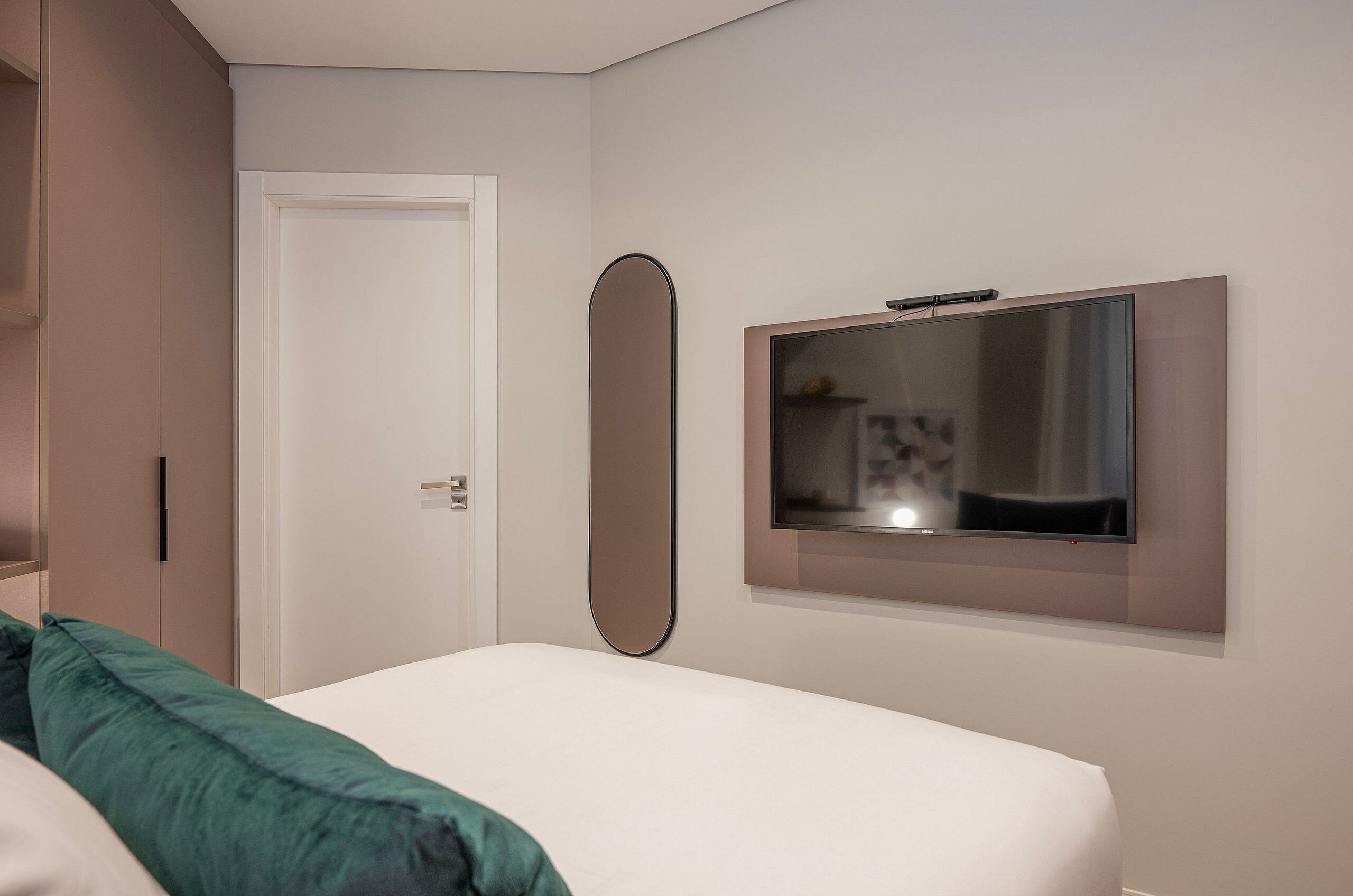 Hotellihuone, jossa on sänky, seinälle asennettu televisio, ovi ja peili.