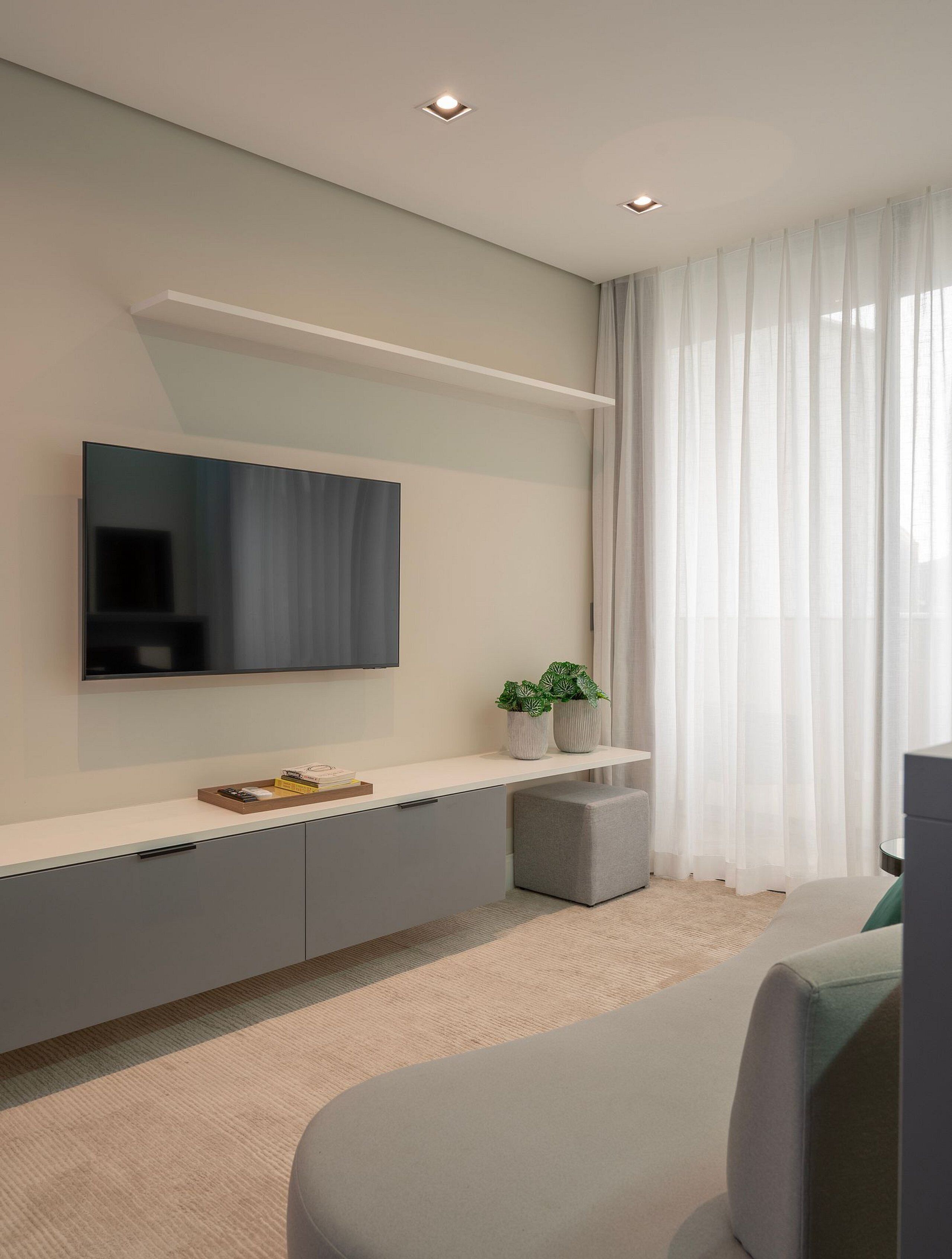 A modern living room with a flat-screen TV mounted on the wall, a grey sofa, a small ottoman, and a white console table with decorative plants.