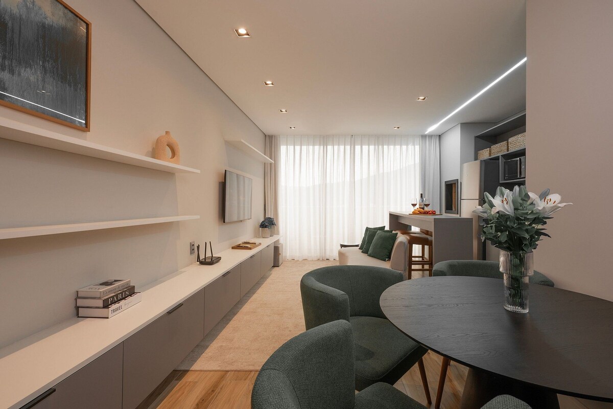 Un salón moderno con comedor, un televisor de pantalla plana montado en la pared y un amplio ventanal con cortinas translúcidas.
