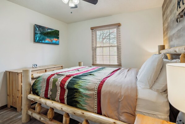 A bedroom with a wooden bed frame, a nightstand, a bedside table, a window with blinds, and a wall-mounted picture. - 3 min to Parkway Tranquil Mtn Hideaway w/ Hot Tub, Arcade, Grill & Outdoor Seats (Pigeon Forge)