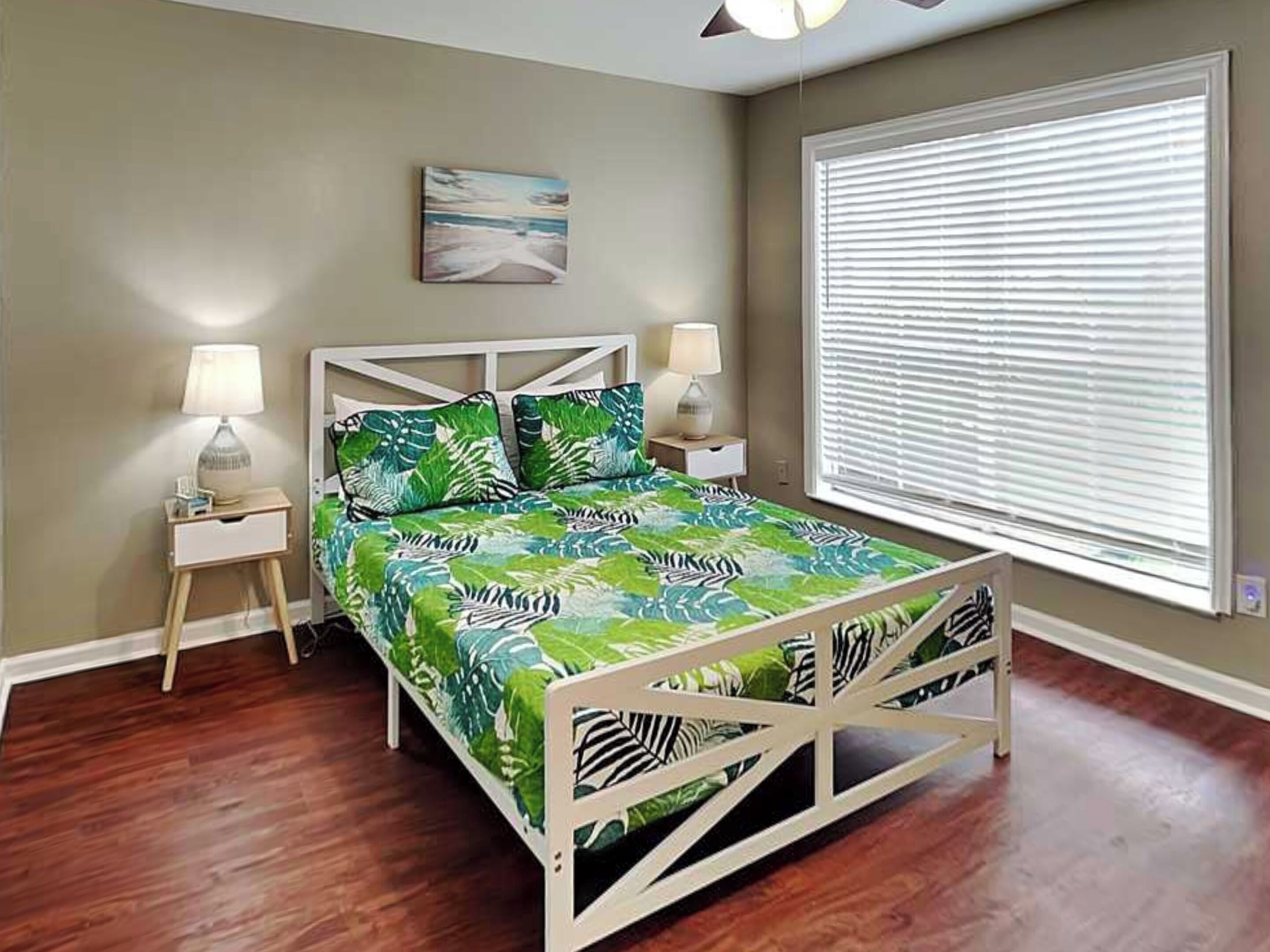 A bedroom with a bed, nightstands, a ceiling fan, a window with blinds, and a wall-mounted artwork.