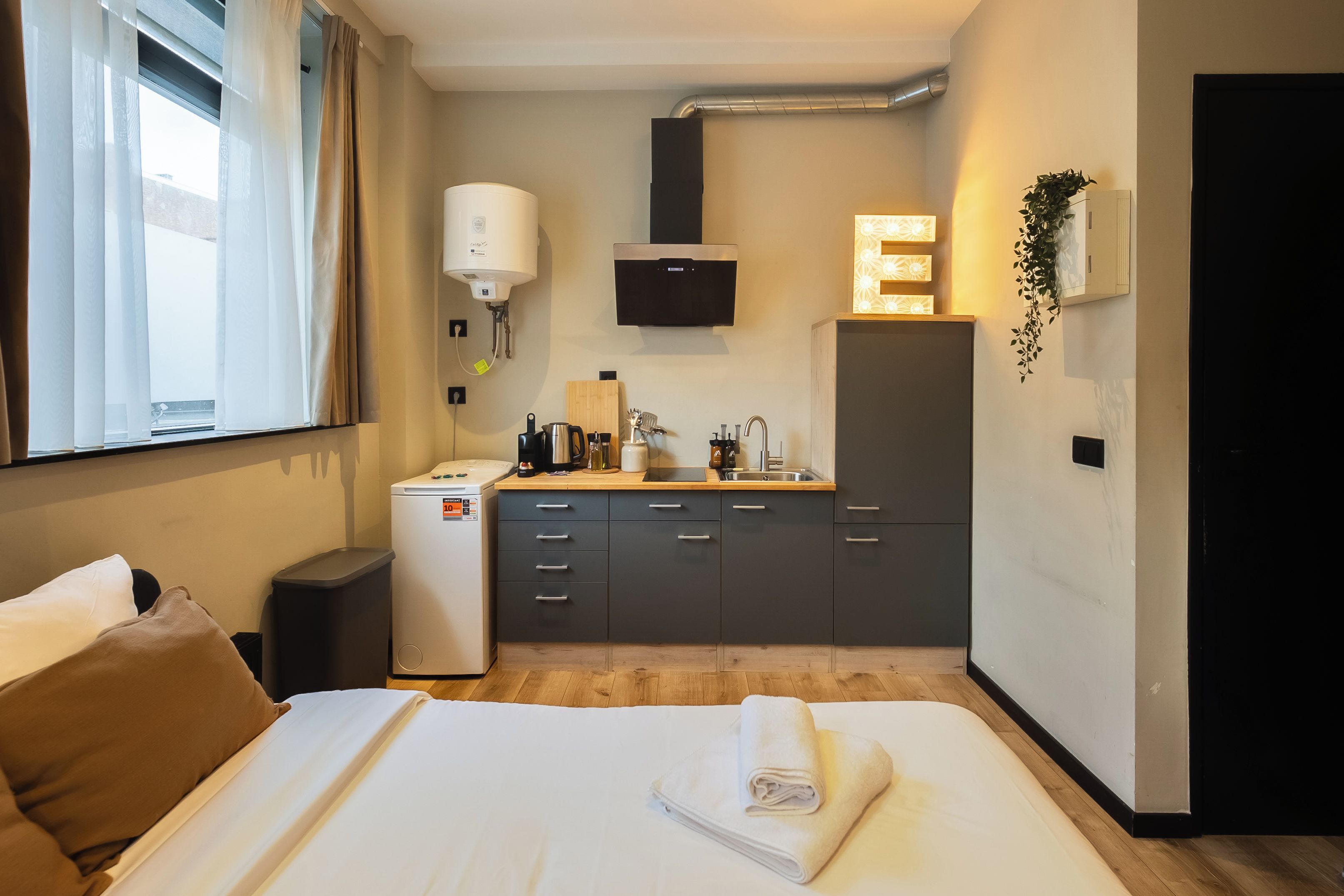 Une chambre d’hôtel compacte comprenant un lit, une petite kitchenette, une fenêtre avec des rideaux et un téléviseur fixé au mur.