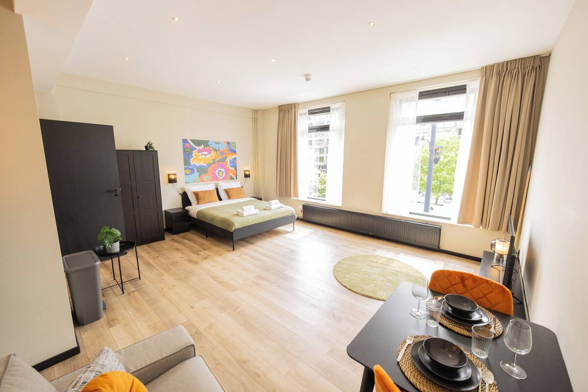 Una habitación de hotel moderna con una cama, una mesa de comedor para dos personas, un sofá y un amplio ventanal con cortinas.