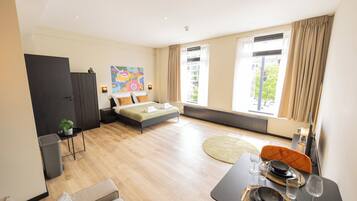 A modern hotel room with a bed, a dining table set for two, a sofa, and a large window with curtains.