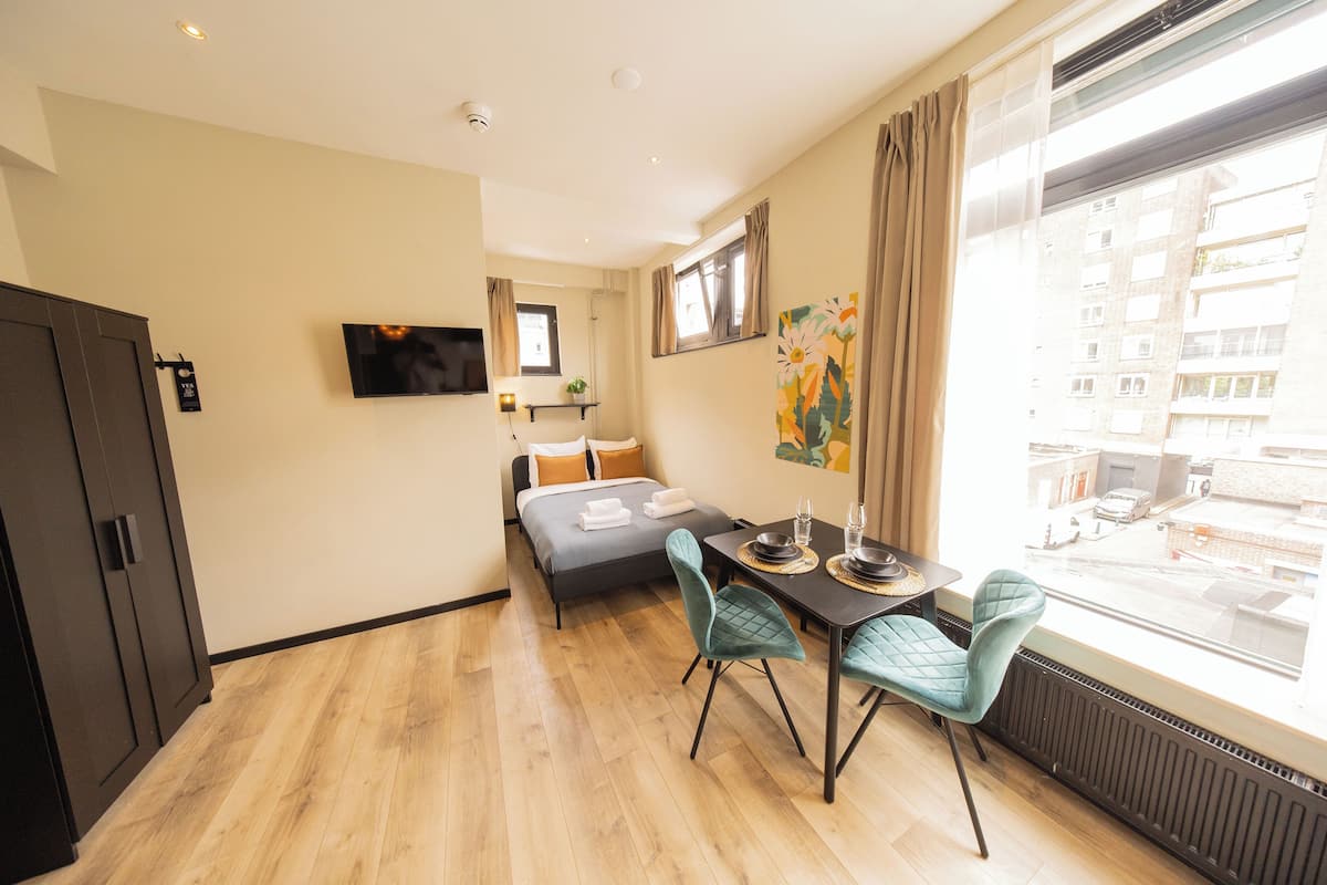 Una habitación de hotel con cama, una mesa de comedor con sillas, un televisor montado en la pared y una ventana con cortinas.