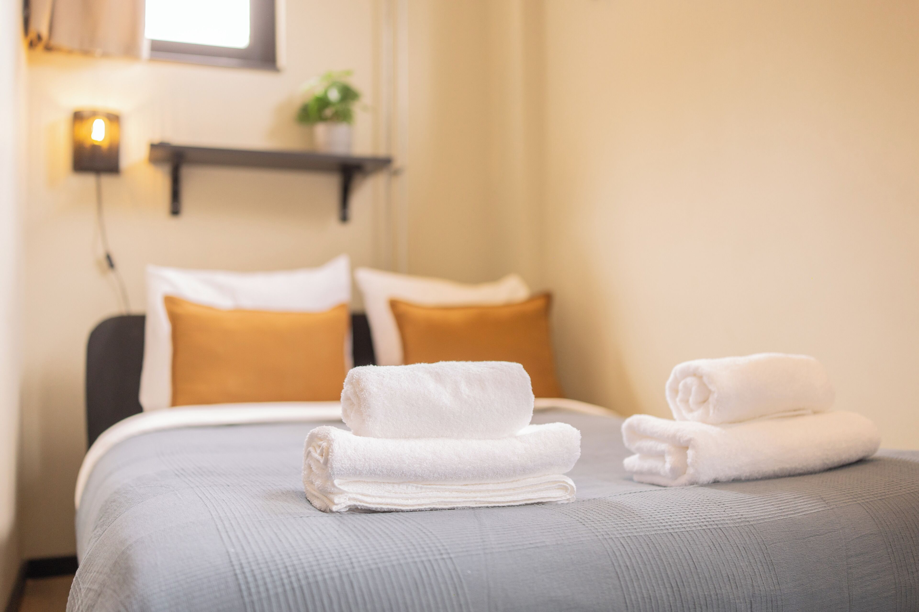 Cama bem arrumada com duas toalhas brancas enroladas e dois travesseiros, um branco e um amarelo mostarda, em um quarto com prateleira na parede e uma pequena planta.