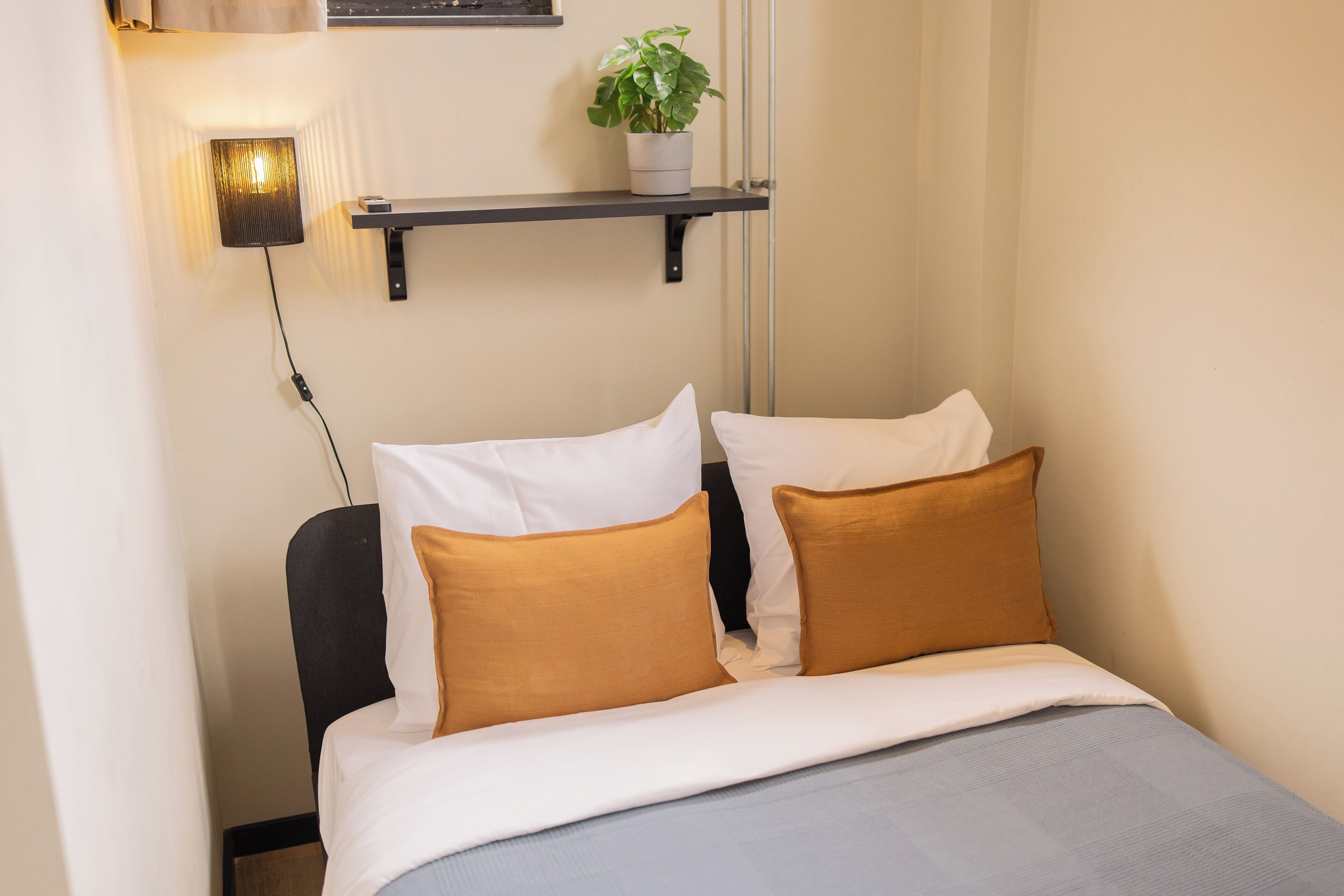 A neatly made bed with white and brown pillows, a grey blanket, and a small potted plant on a shelf above.