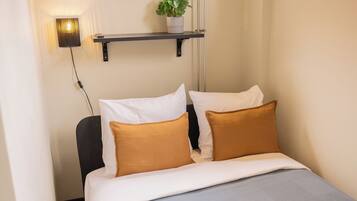 A neatly made bed with white and brown pillows, a grey blanket, and a small potted plant on a shelf above.