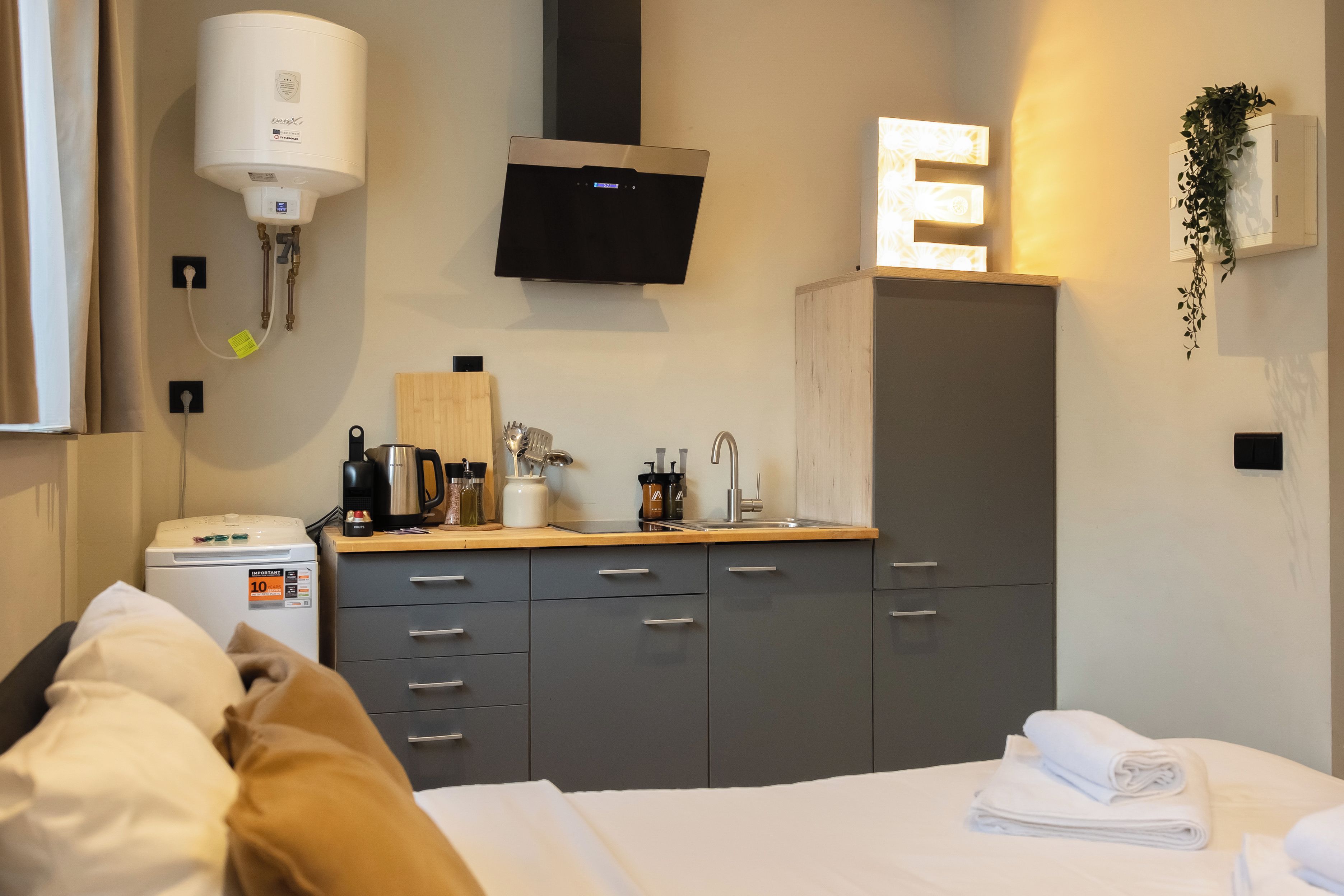 A hotel room with a bed, a desk with a kettle, a coffee maker, and a TV mounted on the wall.
