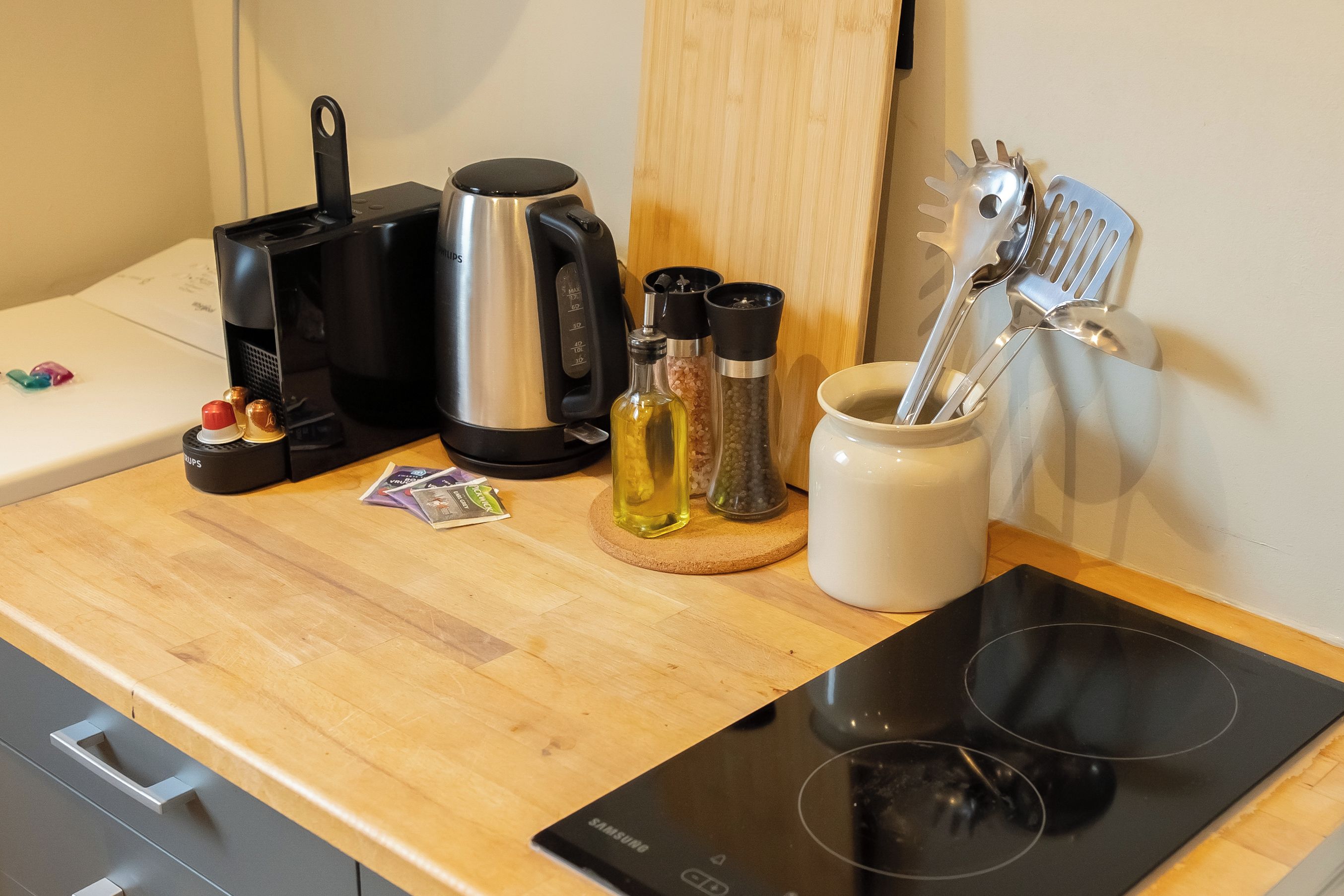 Una encimera de cocina con una cafetera, un hervidor, una botella de aceite, un especiero y un juego de utensilios.