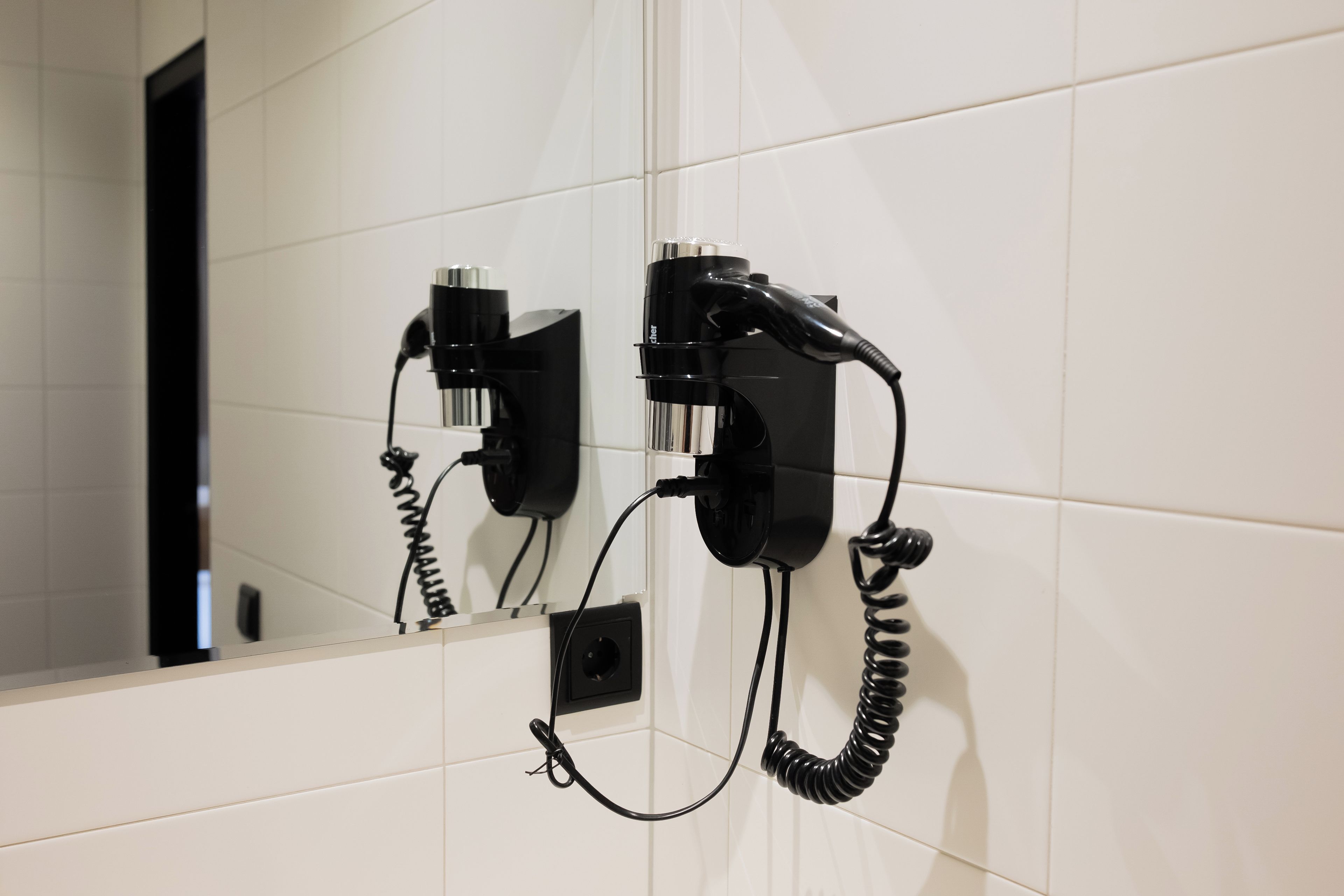 Un sèche-cheveux et un fer à friser, fixés sur un mur recouvert de carreaux blancs, sont reliés par un cordon enroulé.