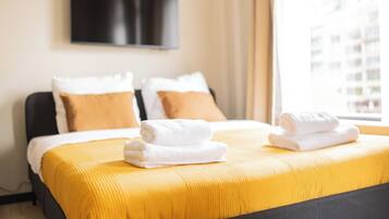 A hotel room with a bed, two pillows, a flat-screen TV on the wall, and a window with a view of buildings.