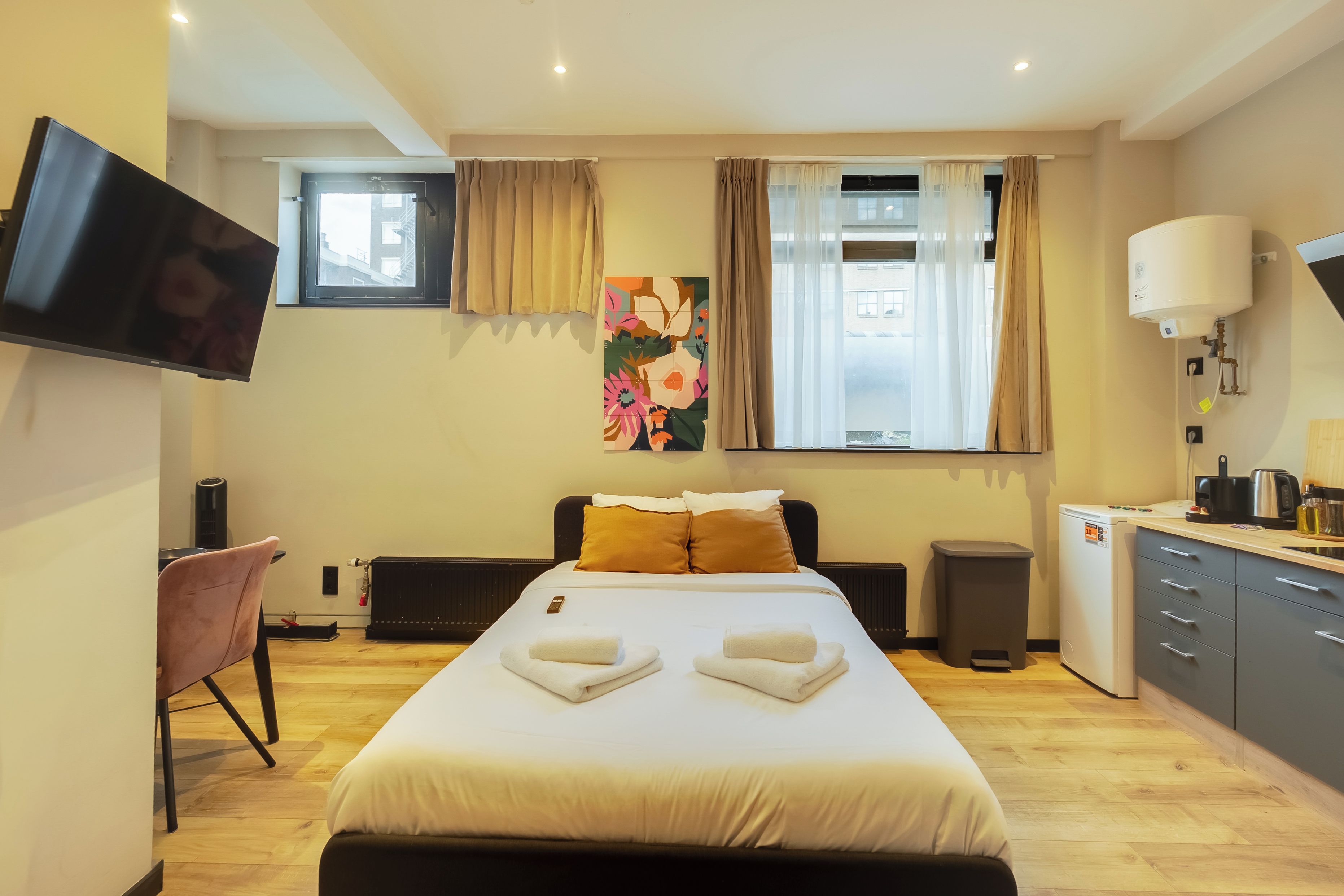 A hotel room with a bed, a desk, a television, a radiator, a window with curtains, and a kitchen area with a kettle and a coffee maker.