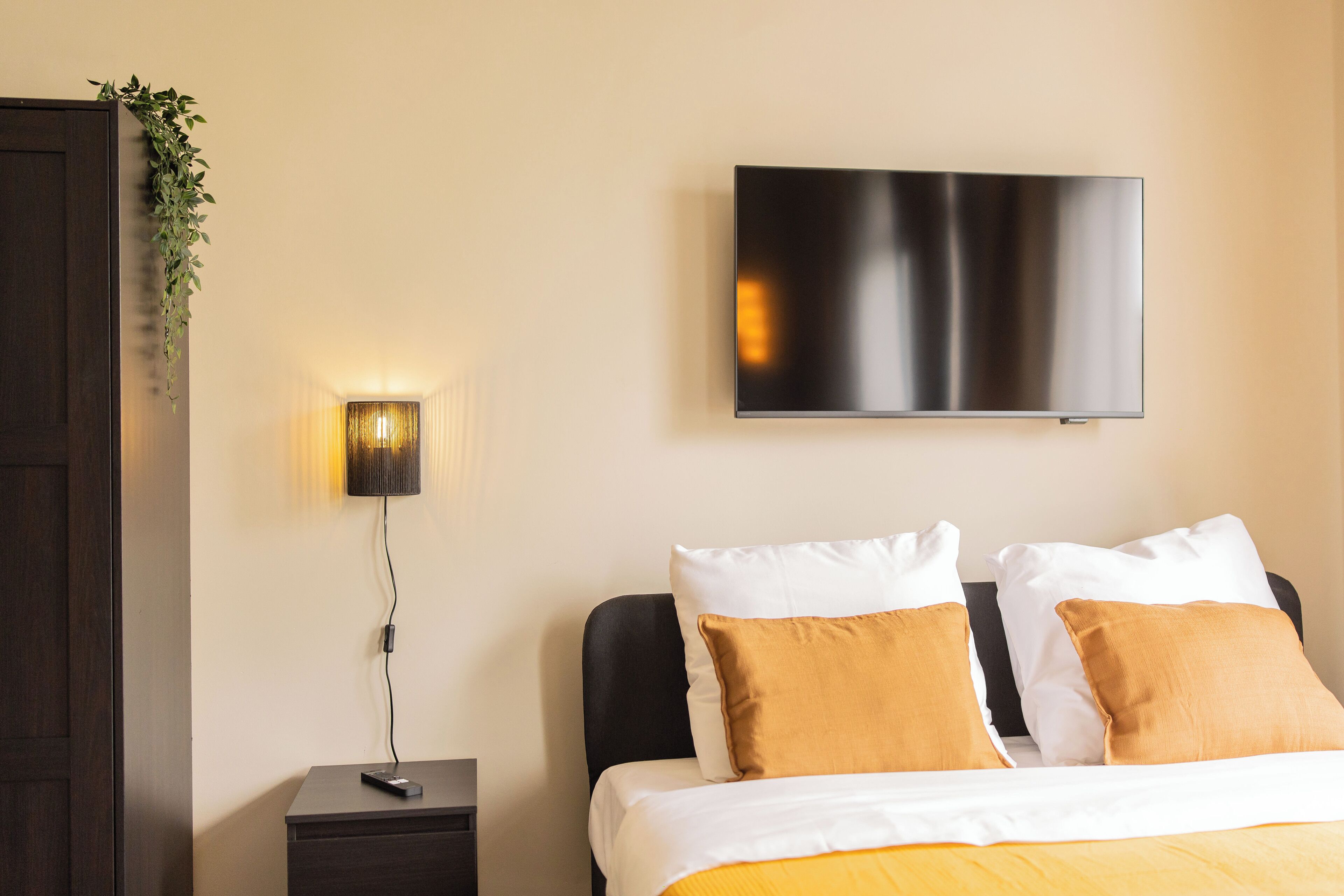 Habitació d'hotel amb un llit, una televisió penjada a la paret, una taula de nit amb una làmpada i una porta de fusta fosca.