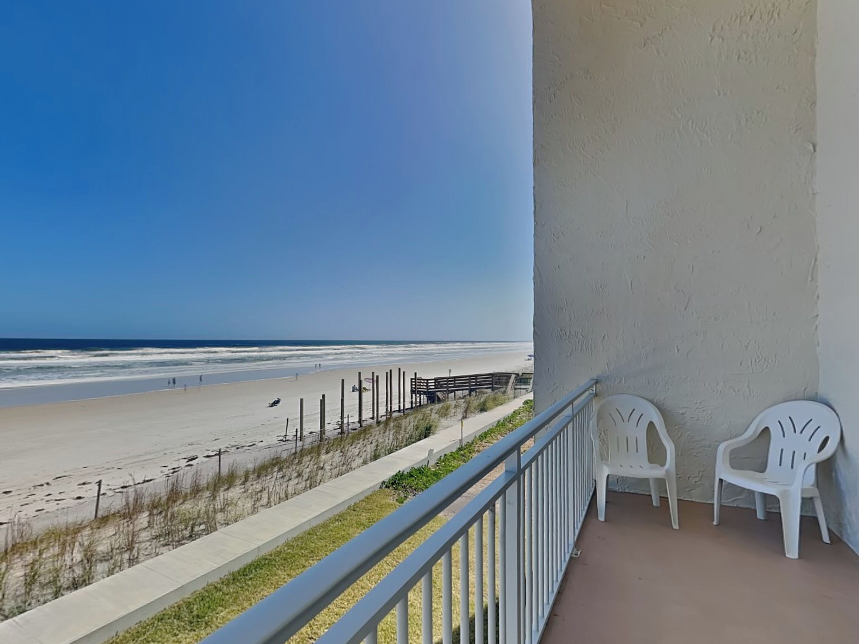 Een balkon met uitzicht op het strand, witte plastic stoelen en een hekwerk.