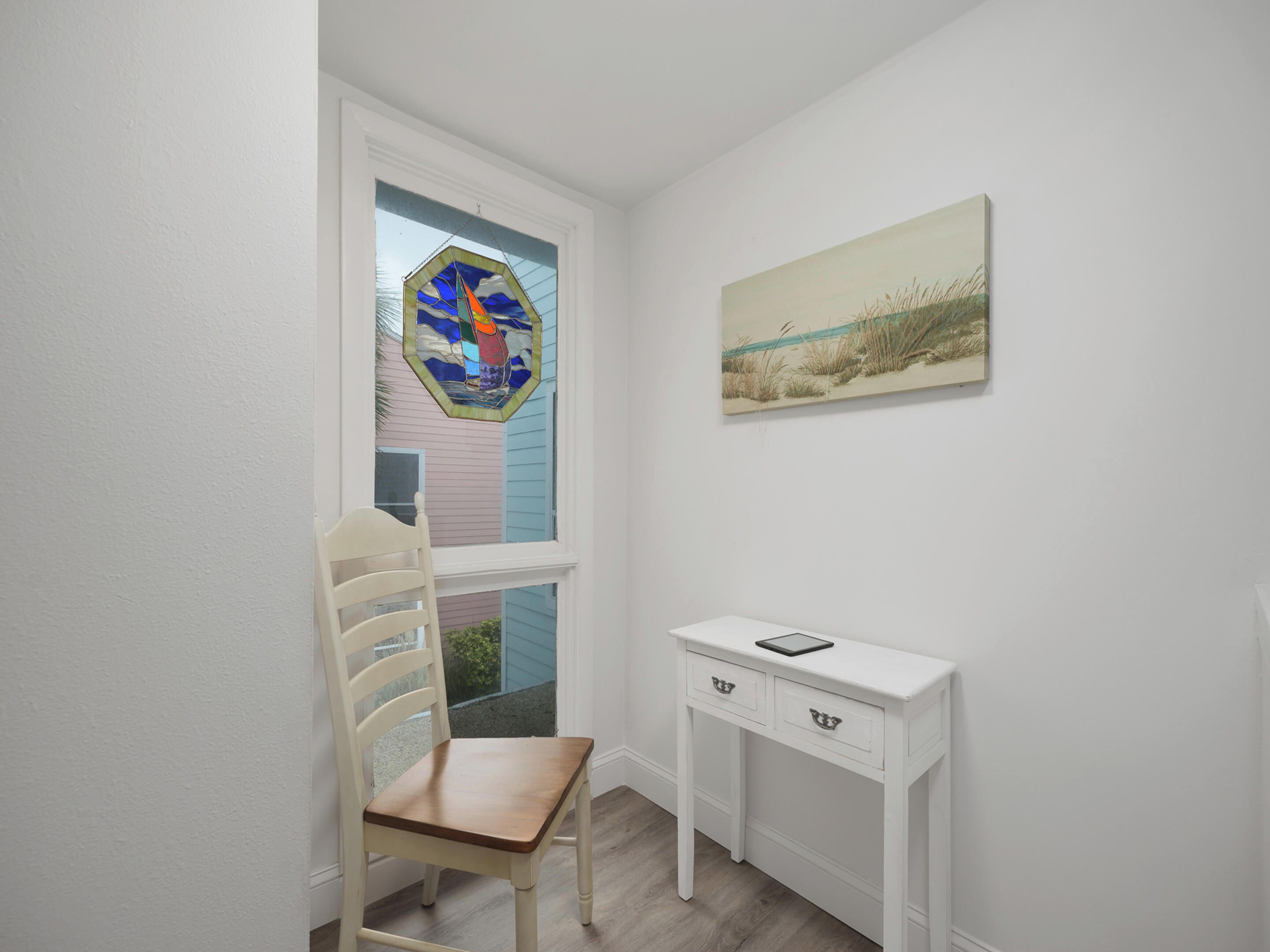 Een kamer met een witte stoel, een wit bureau met lades, een schilderij aan de muur en een glas-in-loodraam.