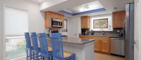 A modern kitchen with a central island, blue chairs, wooden cabinets, and a stainless steel refrigerator.