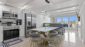 A modern kitchen with stainless steel appliances, a dining area with a white table and cushioned chairs, and a balcony with a view of the sky and palm trees. - Chic Stay by the Beach (South Padre Island)