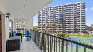 Un balcón con vistas a un edificio residencial alto, mobiliario exterior y una cancha de tenis.