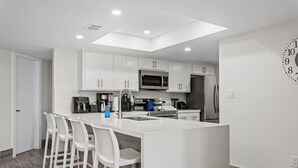 Una cocina moderna con armarios blancos, una isla central con taburetes de bar, un microondas, un refrigerador y un reloj en la pared.