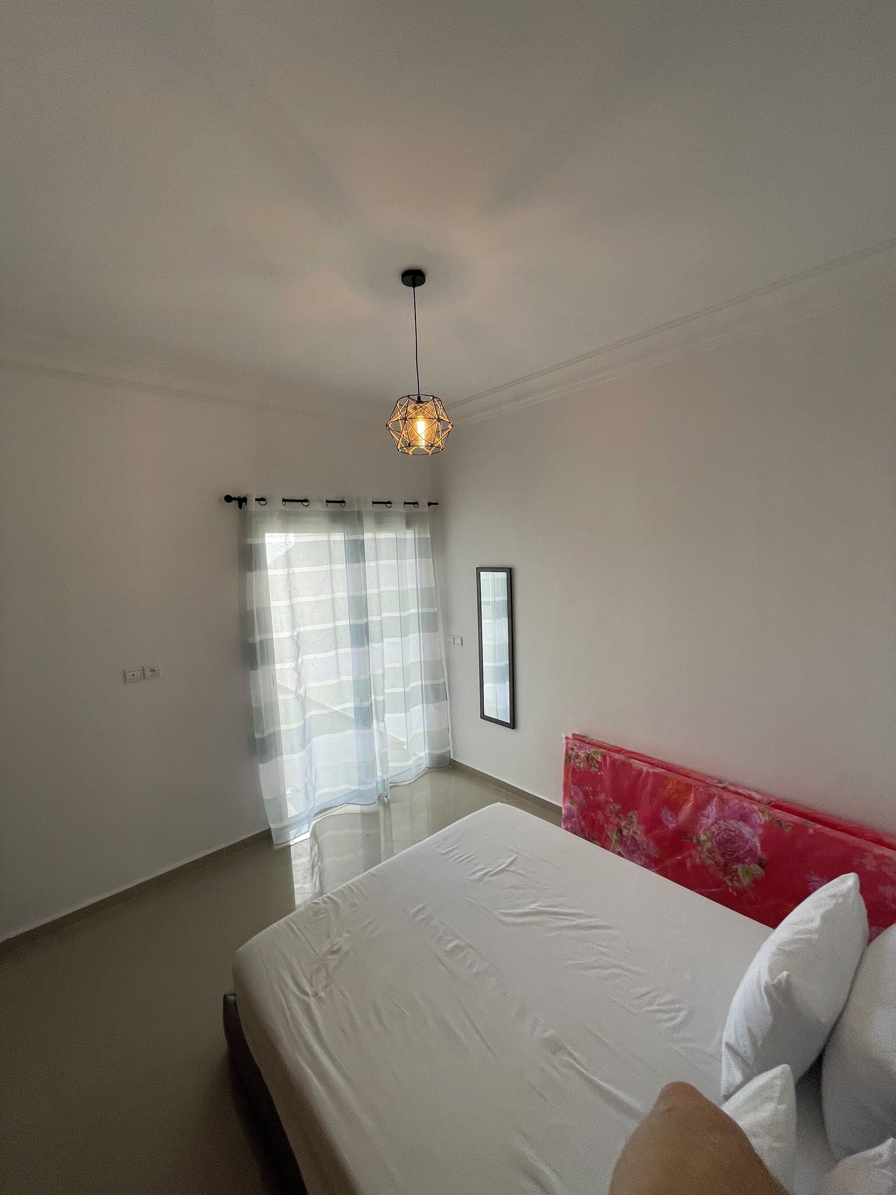 Une chambre à coucher avec un lit, un luminaire suspendu, une fenêtre avec des rideaux légers et un tableau encadré accroché au mur.