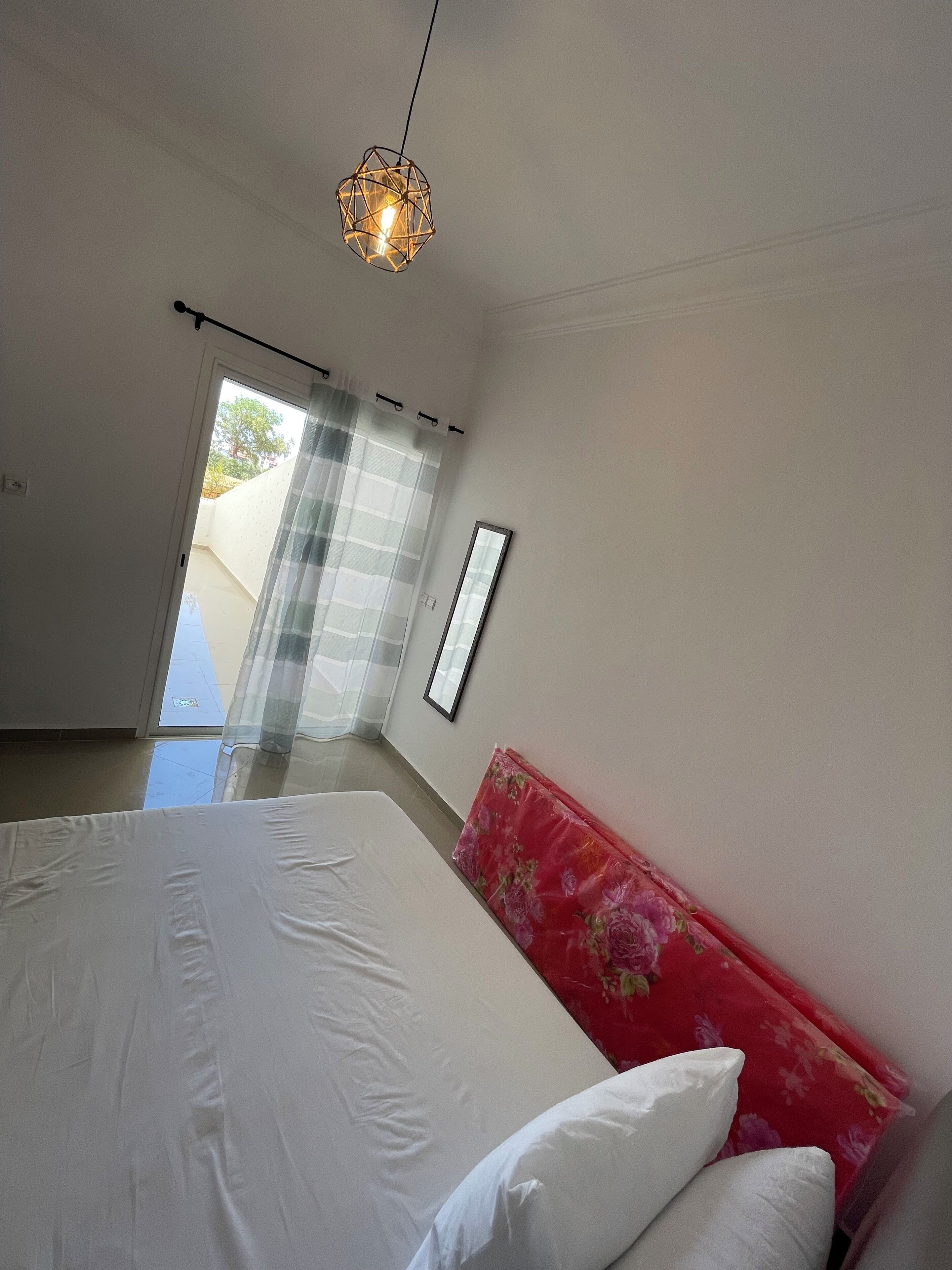 Ein Schlafzimmer mit einem Bett, einer roten Kopfteil, einer Pendelleuchte, einem Fenster mit Vorhängen und einer zur Außenseite führenden Tür.