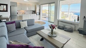 Un salon moderne avec un canapé d’angle gris, une table basse en bois et une vue sur la ville grâce à de grandes fenêtres.