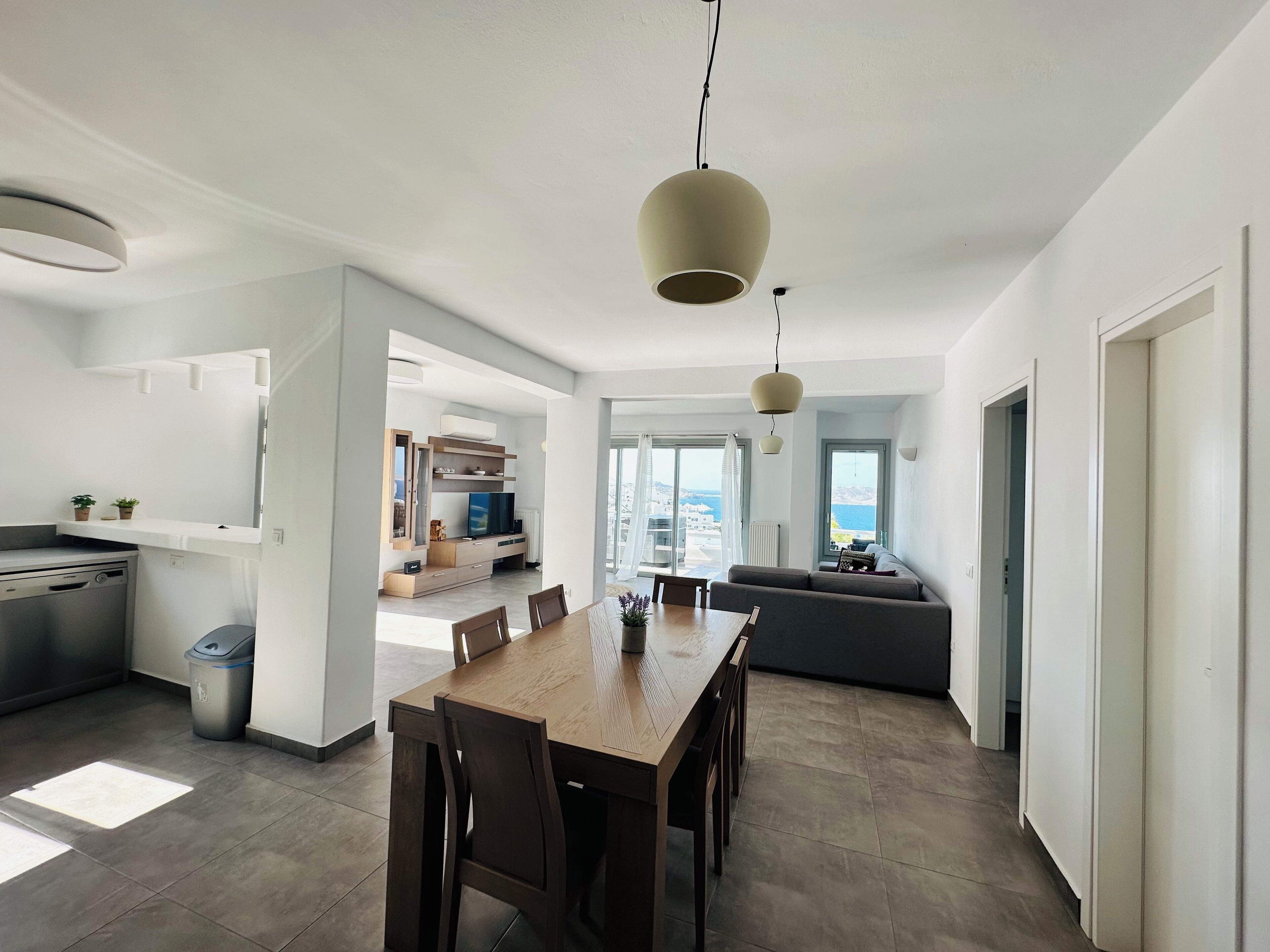 A modern kitchen with a dining area, a grey sofa, and a view of the sea.