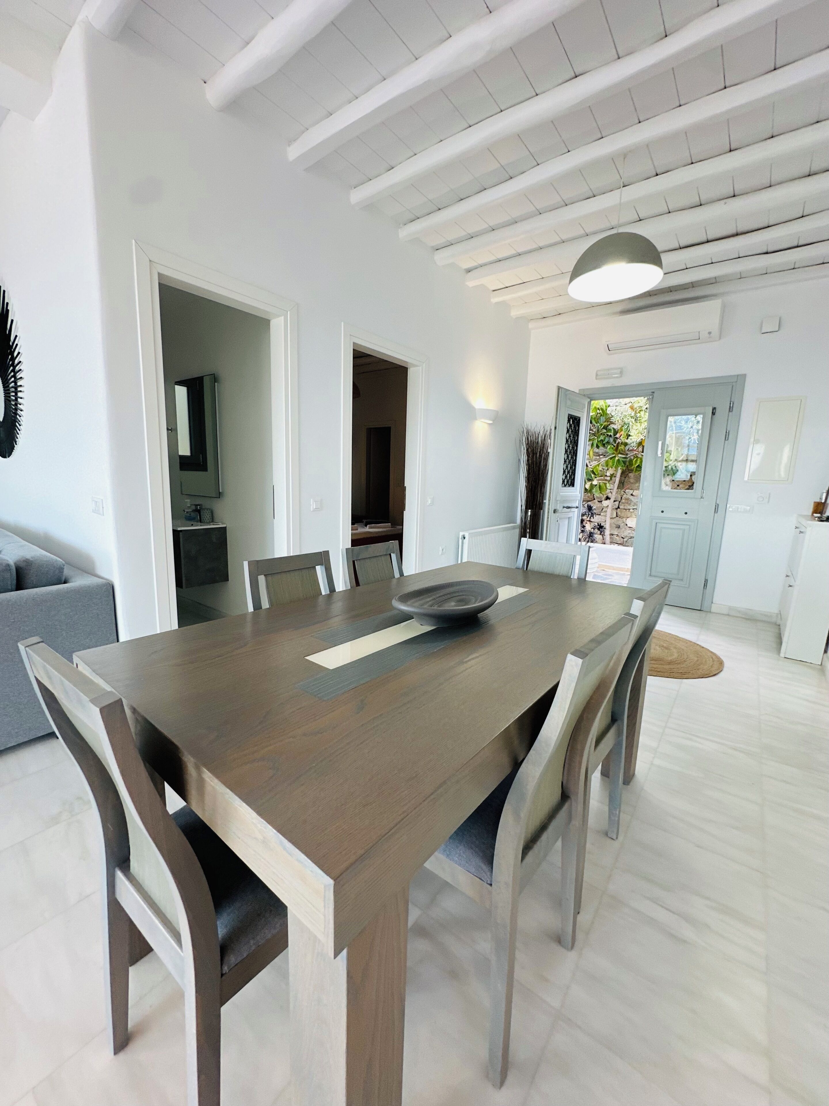 A spacious dining area with a large wooden table and chairs, white walls, and a ceiling with exposed beams.