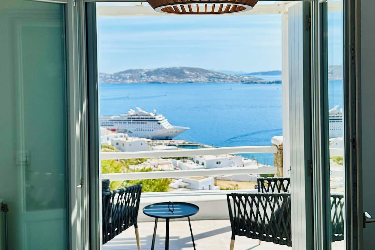 An open balcony with a view of the sea and a cruise ship, featuring a hanging light fixture and two small tables.